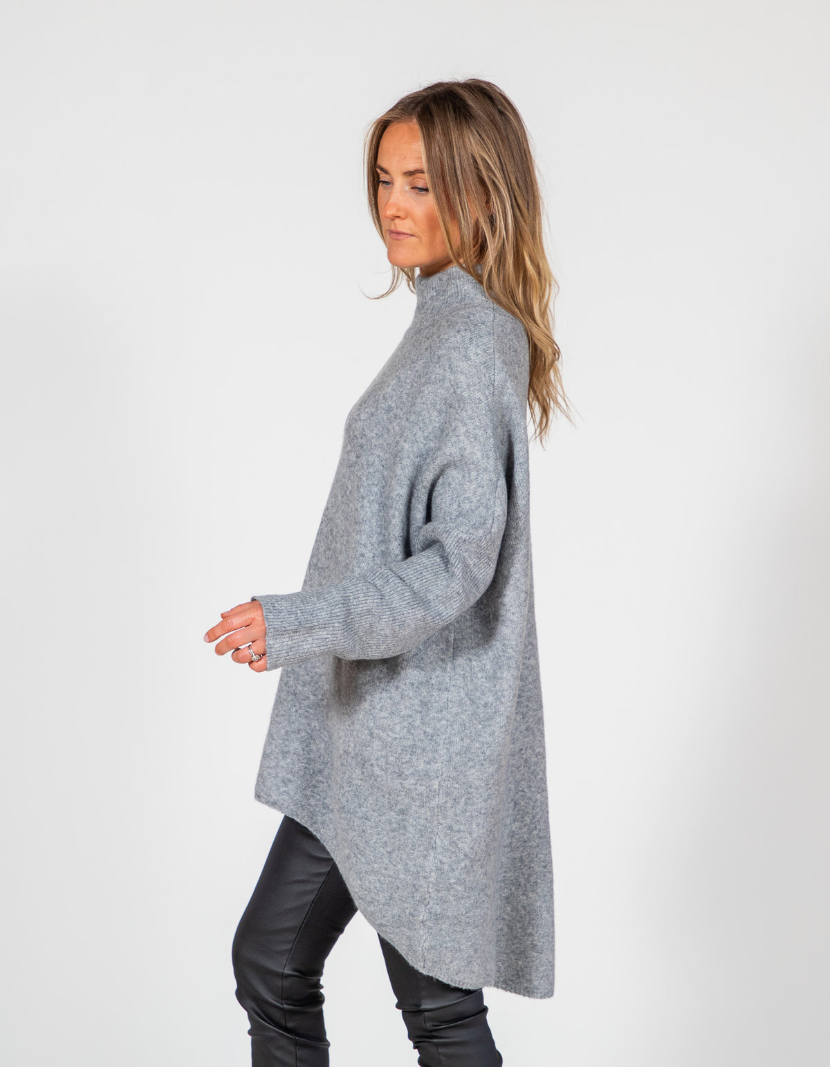 Grey oversized brushed knit jumper seen from the side, featuring a ribbed funnel neck and pronounced high-low hemline.