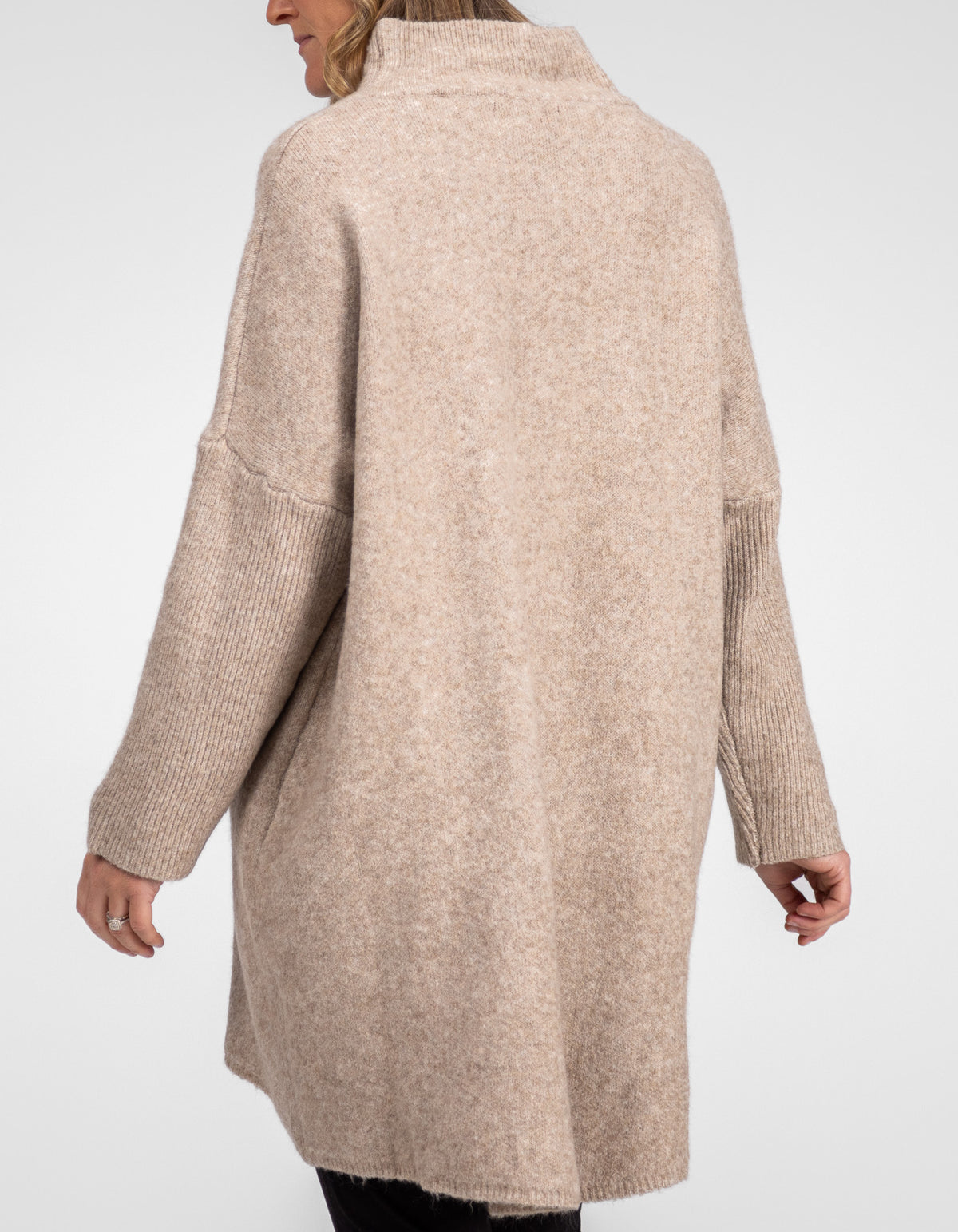 Rear half-body shot showing the high-low hemline and extended back drape of the beige jumper over black pants.